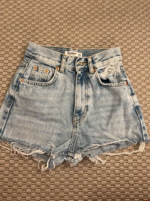 Blåa högmidjade jeansshorts Pull&Bear - Säljer ett par ljusblå högmidjade jeansshorts från Pull&Bear. Shortsen har råa, fransiga kanter och slitningar både fram och bak. Klassisk femficksmodell med knappgylf och bälteshällor. Perfekta för varma dagar!