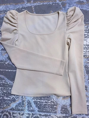 Beige topp med puffärmar - Trendig beige topp med fyrkantig ringning och långa puffärmar. Toppens material känns mjukt och har en lätt struktur, vilket ger en snygg och modern look. Perfekt att styla med jeans eller kjol för en cool outfit.