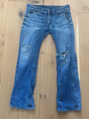 Blå bootcut jeans med slitningar - Säljer ett par blå bootcut jeans från WR med slitna detaljer. Jeansen har en klassisk femficksdesign, ljus tvätt och broderade bakfickor. Perfekta för en avslappnad och trendig look.
