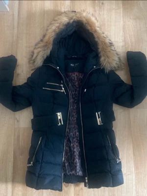 Svart dunjacka med fuskpäls från Hollies - Snygg svart dunjacka från Hollies med avtagbar huva och fuskpäls. Jackan har flera dragkedjefickor, bälte i midjan och leopardmönstrat innerfoder. Perfekt för kalla vinterdagar och riktigt trendig look.