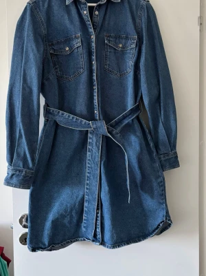 Jeansklänning med bälte från JJXX - Snygg jeansklänning från JJXX i klassisk blå denim. Klänningen har långa ärmar, knappar framtill, två bröstfickor och ett matchande bälte i midjan. Perfekt för en cool och avslappnad look.