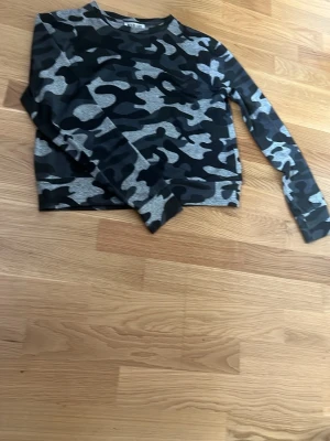 Camo långärmad tröja i grått och svart - Säljer en långärmad tröja med camouflagemönster i grått och svart. Tröjan har rund halsringning och är tillverkad i ett mjukt, stretchigt material. Perfekt för en avslappnad streetstyle-look och funger bra till tränig.