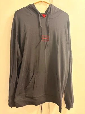 Svart hoodie från Hugo med logga - Svart hoodie från Hugo med röd logotyp på bröstet. Hoodien har huva med snörning och en stor magficka. Tillverkad i mjukt material och har en avslappnad passform med långa ärmar. Perfekt för en chill och stilren look.