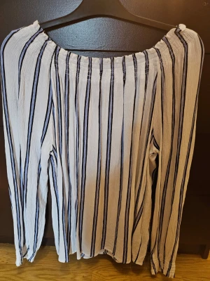 Randig offshoulder blus i vitt och blått - Säljer en snygg offshoulder blus med långa ärmar och vertikala ränder i vitt, blått och svart. Blusen har en luftig och krinklad struktur som ger en avslappnad känsla. Perfekt för dig som gillar trendiga och bekväma plagg. KappAhl 