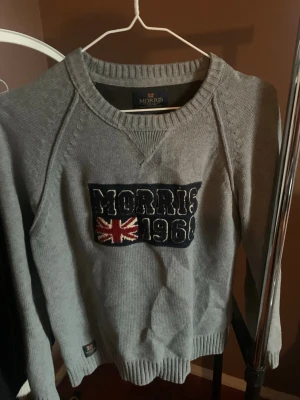 Grå stickad tröja Morris  - Grå stickad tröja från Morris med broderad patch framtill där det står 'Morris 1968' och en brittisk flagga. Tröjan har rund halsringning och ribbade muddar vid ärmslut och nederkant. Perfekt för en avslappnad och stilren look.
