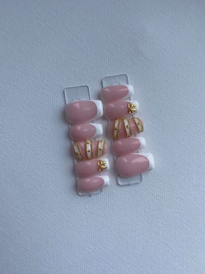 Press-on naglar rosa med gulddetaljer - Snygga press-on naglar i ljusrosa med vit fransk tipp och guldfärgade dekorationer. Några naglar har tunna guldränder och små guldfärgade rosetter för extra bling. Oval form och glansig finish. Perfekt för dig som vill ha en chic och trendig look.