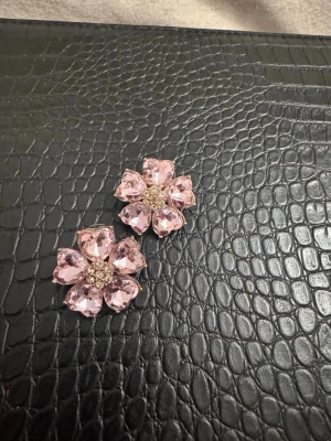 Rosa blomformade örhängen med stenar - Säljer ett par stora örhängen i form av blommor med ljusrosa glittrande stenar som kronblad och små gnistrande stenar i mitten. Örhängena har en lyxig och iögonfallande design, perfekta för att sticka ut. Materialet är troligen metall och glas.