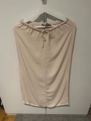 Beige midikjol med snörning från Primark - Stilren beige midikjol från Primark med elastisk midja och snörning framtill. Kjolen har en rak passform och är tillverkad i ett mjukt material som känns skönt mot huden. Perfekt för dig som gillar enkel och clean stil.