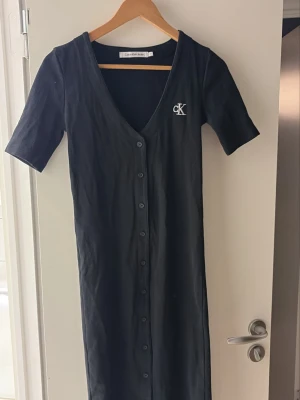 Svart klänning från Calvin Klein Jeans - Svart ribbad klänning från Calvin Klein Jeans med korta ärmar, djup V-ringning och knappar hela vägen framtill. Klänningen har en broderad vit CK-logga på bröstet och är i ett stretchigt material som sitter snyggt på kroppen.