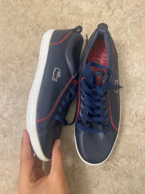 Marinblå Lacoste sneakers med röd detalj - Snygga marinblå sneakers från Lacoste med röda detaljer och vit sula. Skorna har klassisk låg modell, blå snörning och den ikoniska krokodilloggan på sidan. Ovandelen är i slätt syntetmaterial som ger en clean look.