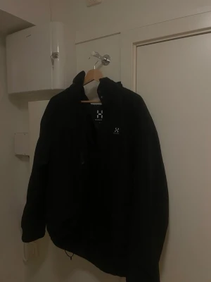 Svart vindjacka från Haglöfs - Snygg svart vindjacka från Haglöfs med hög krage och minimalistisk design. Jackan har dragsko i nederkant och diskret logga på bröstet. Perfekt för blåsiga dagar och enkel att matcha med olika outfits. Trasig dragkedja därav priset.