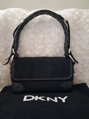 Svart handväska från DKNY - Snygg svart handväska från DKNY med diskret logomönster och detaljer i läder. Väskan har en flätad axelrem med silverfärgade metalldetaljer och klassisk rektangulär form. Perfekt för dig som gillar stilrena accessoarer med en twist.