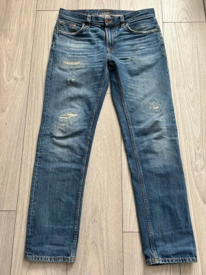 Blå Nudie jeans 31/32 - Säljer ett par blå jeans från Nudie Jeans med snygga slitningar och klassisk femficksmodell. Jeansen har raka ben och orangea sömmar samt detaljerade bakfickor. Vid fundering är det bara att höra av sig!