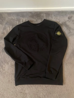 Svart sweatshirt från Stone Island - Svart sweatshirt från Stone Island med broderad logga på bröstet och klassisk patch på vänster ärm. Tröjan har rund hals och ribbade muddar. Materialet är mjuk bomull och designen är stilren med streetwear-vibe.