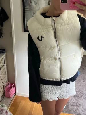 Vit dunväst från True Religion - Snygg vit dunväst från True Religion med dragkedja framtill och hög krage. Västen har en svart logga på bröstet och stor svart text längs sidan. Perfekt för lager-på-lager och streetstyle. Materialet är glansigt och quiltat för extra värme.