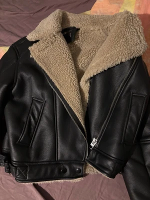 Svart teddyjacka från FB Sister XS - Snygg svart teddyjacka från FB Sister i storlek XS. Jackan har mjukt beige teddyfoder, bred krage och detaljer i fuskskinn. Dragkedja framtill och två fickor med snedställd öppning. Perfekt för kalla dagar och ger en cool look.