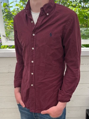 Vinröd skjorta från Ralph Lauren - Säljer en vinröd skjorta från Ralph Lauren med klassisk krage och knappar framtill. Skjortan har lång ärm och den ikoniska loggan broderad på bröstet. Perfekt för dig som gillar stilrena plagg med snygg passform.