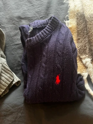 Mörkblå kabelstickad tröja Polo Ralph Lauren - Kabelstickad mörkblå tröja från Polo Ralph Lauren med klassisk röd logga broderad på bröstet. Tröjan har rund halsringning och ribbade muddar. Perfekt för dig som gillar tidlös och preppy stil.