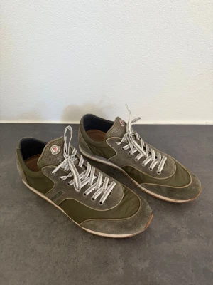 Olivgröna sneakers från Moncler stl 44 - Snygga olivgröna sneakers från Moncler med vita snören och detaljer i mocka och textil. Skorna har låg profil och klassisk Moncler-logga på plösen. Perfekta för dig som gillar stilrena och sportiga skor med exklusiv känsla.