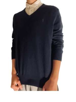 Mörkblå V-ringad tröja Polo Ralph Lauren - Klassisk mörkblå V-ringad tröja från Polo Ralph Lauren. Tröjan har lång ärm, diskret logga på bröstet och ribbade muddar. Perfekt att bära över en skjorta för en clean och stilren look. Passar perfekt nu till golfen i våren