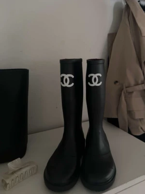 Svarta gummistövlar från Chanel - Snygga svarta gummistövlar från Chanel med det klassiska vita CC-loggan framtill på skaftet. Stövlarna är höga och har en rund tå samt mönstrad sula med logotyp. Perfekta för regniga dagar och en trendig look.