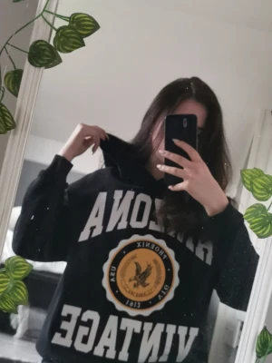 Svart hoodie Arizona Vintage - Cool svart hoodie med stor vit text 'ARIZONA VINTAGE' och ett gult emblem med örn och texten 'PHOENIX USA 1912' på bröstet. Hoodien har huva och en avslappnad passform, perfekt för en chill och trendig look.