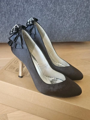Svarta pumps med rosett från Skopunkten - Stilrena svarta pumps i satinliknande material från Skopunkten. Skorna har spetsig tå, smal klack i metallic och dekorativa rosetter med nitar baktill. Perfekta för dig som vill ha en elegant och trendig look. Storlek 36, klackhöjd 8 cm.