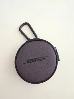 Svart hörlurar från Bose - Säljer en svarta hörlurar med väska från Bose med dragkedja och karbinhake. Perfekt för att förvara småprylar som hörlurar eller mynt. Väskan är tillverkad i slitstarkt tygmaterial och har Bose-loggan tryckt på framsidan.