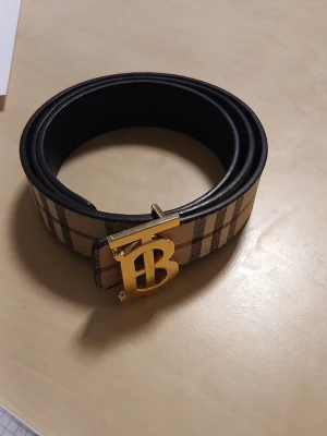 Burberry bälte med rutigt mönster - Snyggt bälte från Burberry med klassiskt beige och brunt rutigt mönster och en guldig spänne med Burberrys logotyp. Bältet har svarta kanter och insida, perfekt för att lyfta vilken outfit som helst.