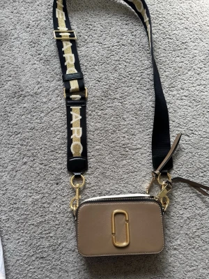 Marc Jacobs Snapshot axelväska beige - Trendig beige axelväska från Marc Jacobs med justerbar svart axelrem med logga i gult och vitt. Väskan har guldfärgade metalldetaljer, dubbla dragkedjor och en praktisk ficka på baksidan. Tillverkad i strukturerat läder med stilren, rektangulär form.