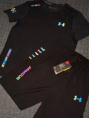 Svart sommarsset från Under Armour - Snyggt svart sommarsset från Under Armour med t-shirt och shorts. Setet har färgglada loggor och detaljer i regnbågsfärger på både tröja och byxa. Materialet är lätt och andas, perfekt för träning eller sportaktiviteter. Modern passform och cool design.