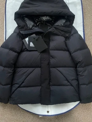 Svart dunjacka med huva från Moncler - Snygg svart dunjacka från Moncler med stor huva och quiltad design. Jackan har ribbade muddar, diskret logotyp på ärmen och dragkedja med knappslå. Perfekt för kalla vinterdagar och tillverkad i ett glansigt material som ger en exklusiv känsla.