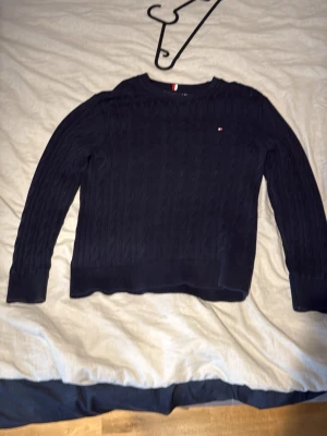 Mörkblå kabelstickad tröja Tommy Hilfiger - Snygg mörkblå kabelstickad tröja från Tommy Hilfiger med rund halsringning och diskret logga på bröstet. Tröjan har ribbade muddar vid ärmslut och nederkant, perfekt för en stilren och klassisk look.