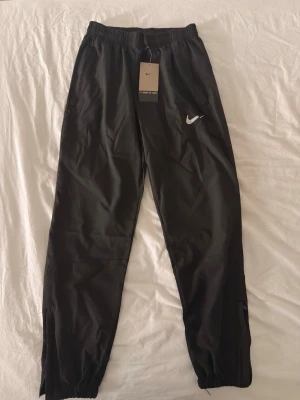 Svarta träningsbyxor från Nike - Svarta träningsbyxor från Nike med elastisk midja och resår vid benslut. Byxorna har en vit Nike-logga på vänster lår och är tillverkade i ett lätt och smidigt material som passar perfekt för sport och träning.