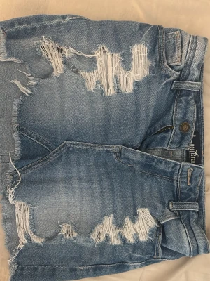 Jeanskjol från hollister - Snygga blå jeansshorts från Hollister med slitna detaljer och fransig kant. Klassisk femficksmodell med knapp och dragkedja. Perfekta för en avslappnad och trendig sommarstil.