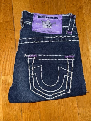 True Religion mörkblå jeans Super T - Snygga True Religion jeans i mörkblå tvätt med klassiska kraftiga kontrastsömmar och lila detaljer. Modell: Stelle Super T – smal passform med stretch för en bekväm och följsam fit.  Premium denim med ikoniska bakfickor och tydlig True Religion-look.  Storlek: 26 Midja: ca 35 cm (platt mätt) Innerbenslängd: ca 80 cm  Material: bomull med stretch (sitter snyggt och bekvämt)  Skick: Nya utan prislapp