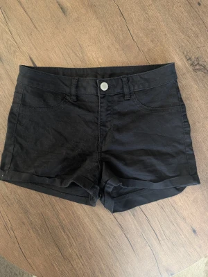 Svarta shorts i bomull - Svarta klassiska shorts med uppvikta benslut och knappstängning framtill. Modellen har fickor fram och bak, och är sydd i ett mjukt bomullstyg. Perfekta för varma dagar och enkel att matcha med olika toppar.