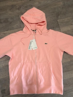 Ljusrosa hoodie från Lacoste - Snygg ljusrosa hoodie från Lacoste med dragkedja framtill, huva med dragsko och klassisk krokodil-logga på bröstet. Mjuk bomullskänsla och två stora fickor framtill. Perfekt för en chill och stilren look.