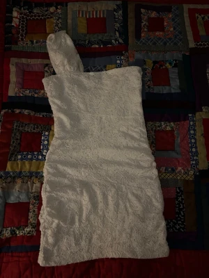 Vit spetsklänning oneshoulder - Snygg vit kort klänning med spets och oneshoulder-design. Klänningen har en figurnära passform och är perfekt för dig som vill sticka ut. Spetsmaterialet ger en romantisk känsla och den asymmetriska axeln gör looken unik.