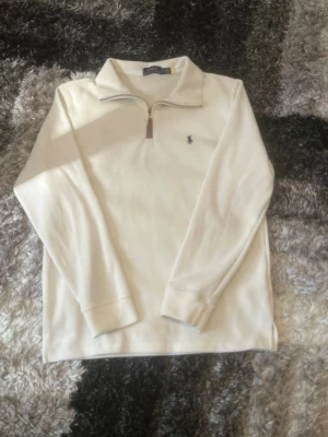 Vit quarter zip från Polo Ralph Lauren - Vit långärmad tröja med quarter zip och liten broderad logga på bröstet från Polo Ralph Lauren. Tröjan har hög krage och ribbade muddar. Perfekt för en clean och stilren look.