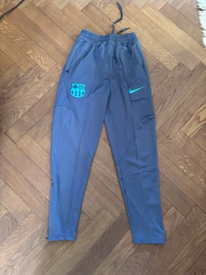 Barça träningsbyxor från Nike - Grå träningsbyxor från Nike med FC Barcelona-logga och turkos Nike-swoosh. Byxorna har dragkedjor vid bensluten, resår i midjan och praktiska fickor. Perfekta för fotbollsträning eller chill. Materialet är lätt och andas.