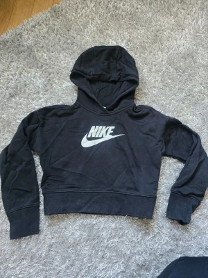 Svart cropped hoodie från Nike 128-134 - Svart cropped hoodie från Nike med stor vit logga på bröstet. Tröjan har huva och långa ärmar, tillverkad i mjukt bomullsmaterial. Perfekt för en sportig och avslappnad stil. Hund finns I hem