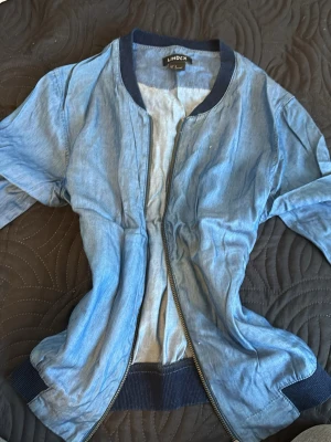 Ljusblå bomberjacka från Lindex - Snygg ljusblå bomberjacka från Lindex i denimliknande material. Jackan har svarta muddar vid ärmslut, krage och nederkant samt dragkedja framtill. Perfekt för en avslappnad och trendig look.