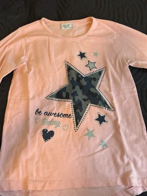 Ljusrosa långärmad topp med stjärnor - Ljusrosa långärmad topp från Name It med stort stjärntryck i camouflage och flera mindre stjärnor framtill. Texten 'be awesome today' och hjärtdetalj. Mjuk bomullskvalitet och rund halsringning.