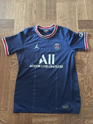PSG matchtröja blå Jordan x Nike strl: 147-158  - Snygg blå Paris Saint-Germain matchtröja med Jordan-logga och klubbmärke på bröstet. Tröjan har vita och röda detaljer på krage och ärmslut samt sponsortryck framtill. Tillverkad i lätt och ventilerande material, perfekt för fotbollsträning.
