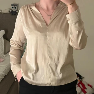 Beige v-ringad blus med långa ärmar - Stilren beige blus med v-ringning och långa ärmar. Blusen har breda manschetter med knappar och är tillverkad i ett lätt, glansigt material som ger en elegant känsla. Perfekt att matcha med både jeans och kostymbyxor.