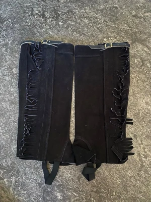 Svarta cowboy boots med fransar - Snygga svarta cowboy boots i mocka med dekorativa fransar längs sidorna. Bootsens höga skaft har spännen upptill och förstärkning vid vristen. Perfekta för dig som vill sticka ut med westernstil.