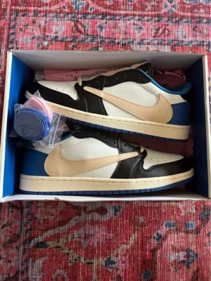 Nike x Travis Scott Jordan 1 Low sneakers - Säljer ett par Nike x Travis Scott Jordan 1 Low sneakers med beige swoosh, svart och vit ovandel samt blå häl. Skorna har snörning och levereras med extra skosnören i olika färger. Perfekt för dig som vill sticka ut med en ikonisk sneaker.