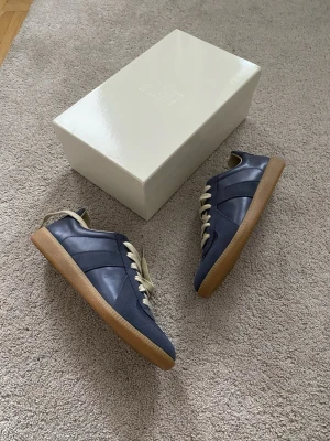 Mörkblå Maison Margiela sneakers - Snygga mörkblå sneakers från Maison Margiela med klassisk design. Skorna har ovandel i läder och mocka, beige snörning och en gummisula i ljusbrun färg. Perfekta för dig som gillar stilrena och tidlösa sneakers med en lyxig touch. Äkthetsverifierade maison margiela gats.