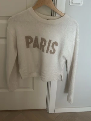 Vit stickad tröja med PARIS-tryck - Vit stickad tröja med beige texten PARIS framtill. Tröjan har rund halsringning, långa ärmar och ribbstickade muddar vid ärmslut och nederkant. Mjuk och lite croppad modell som passar perfekt till jeans eller kjol.